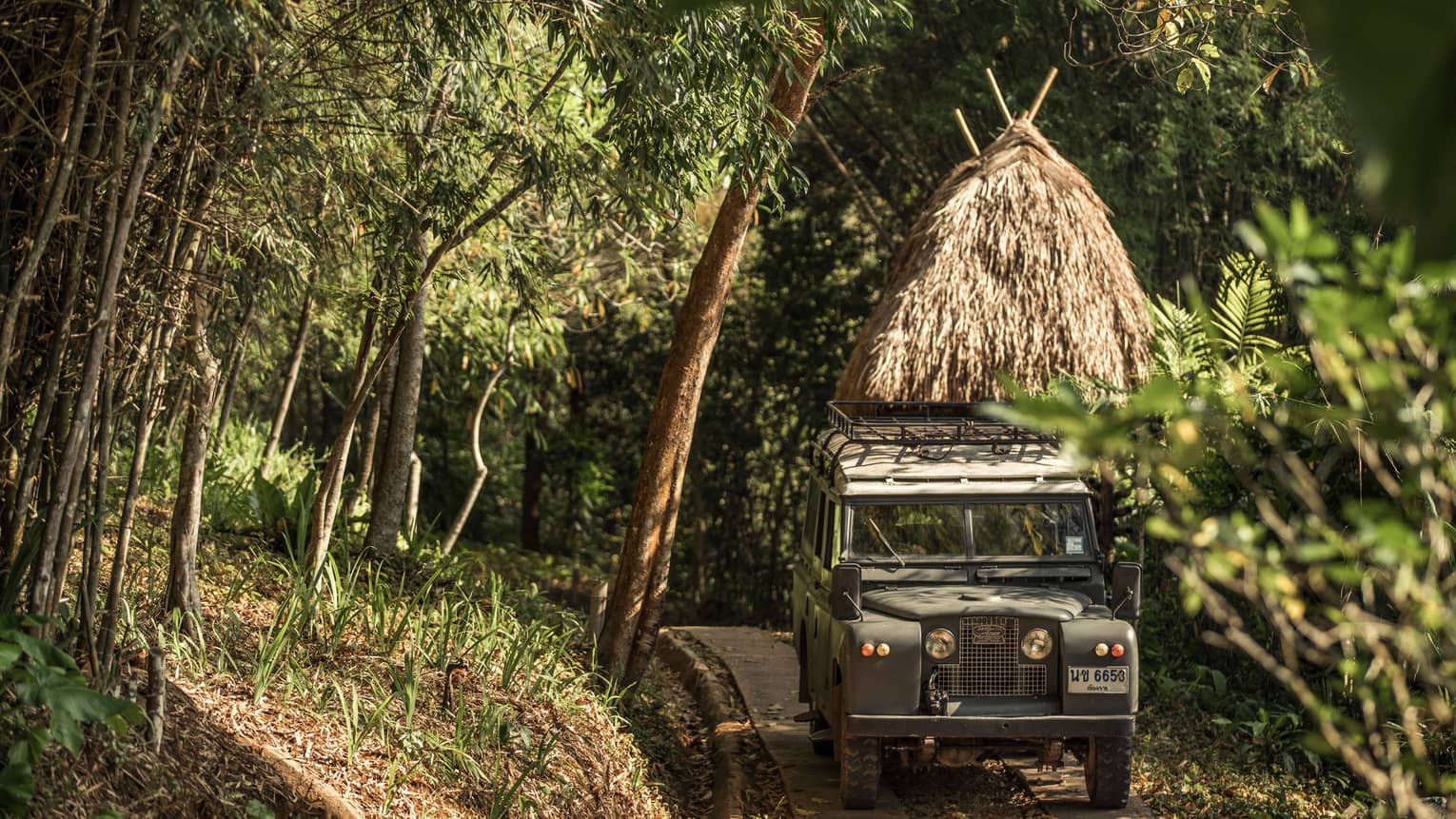 Jeep driving down path in forest past thatched-roof hut