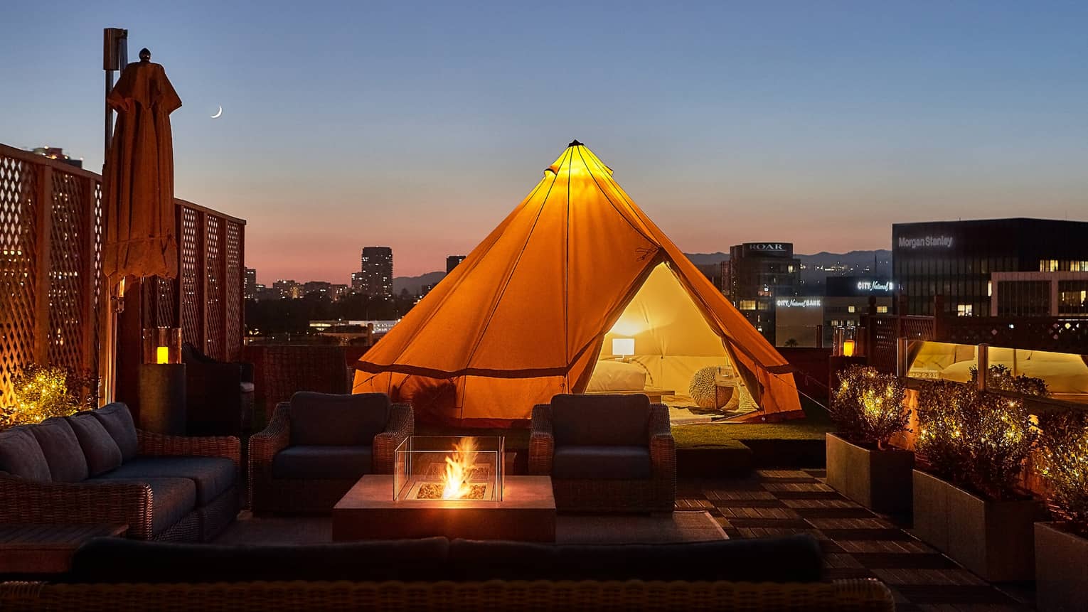Close-up of illuminated white tent with bed, lamp by wicker chairs, modern outdoor fireplace