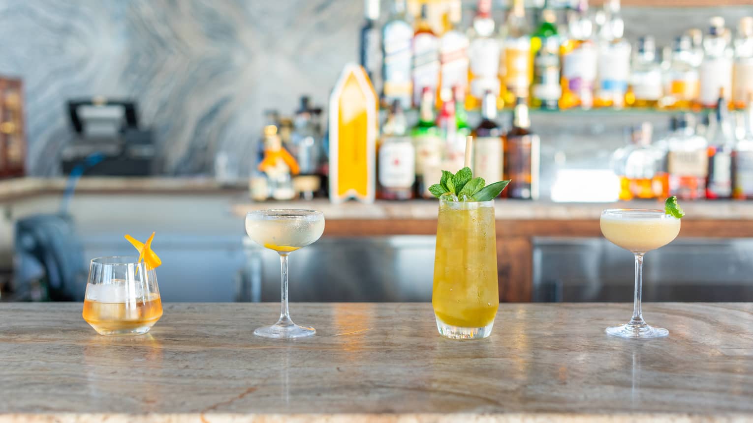 Four cocktails on a bar counter with bottles behind them.
