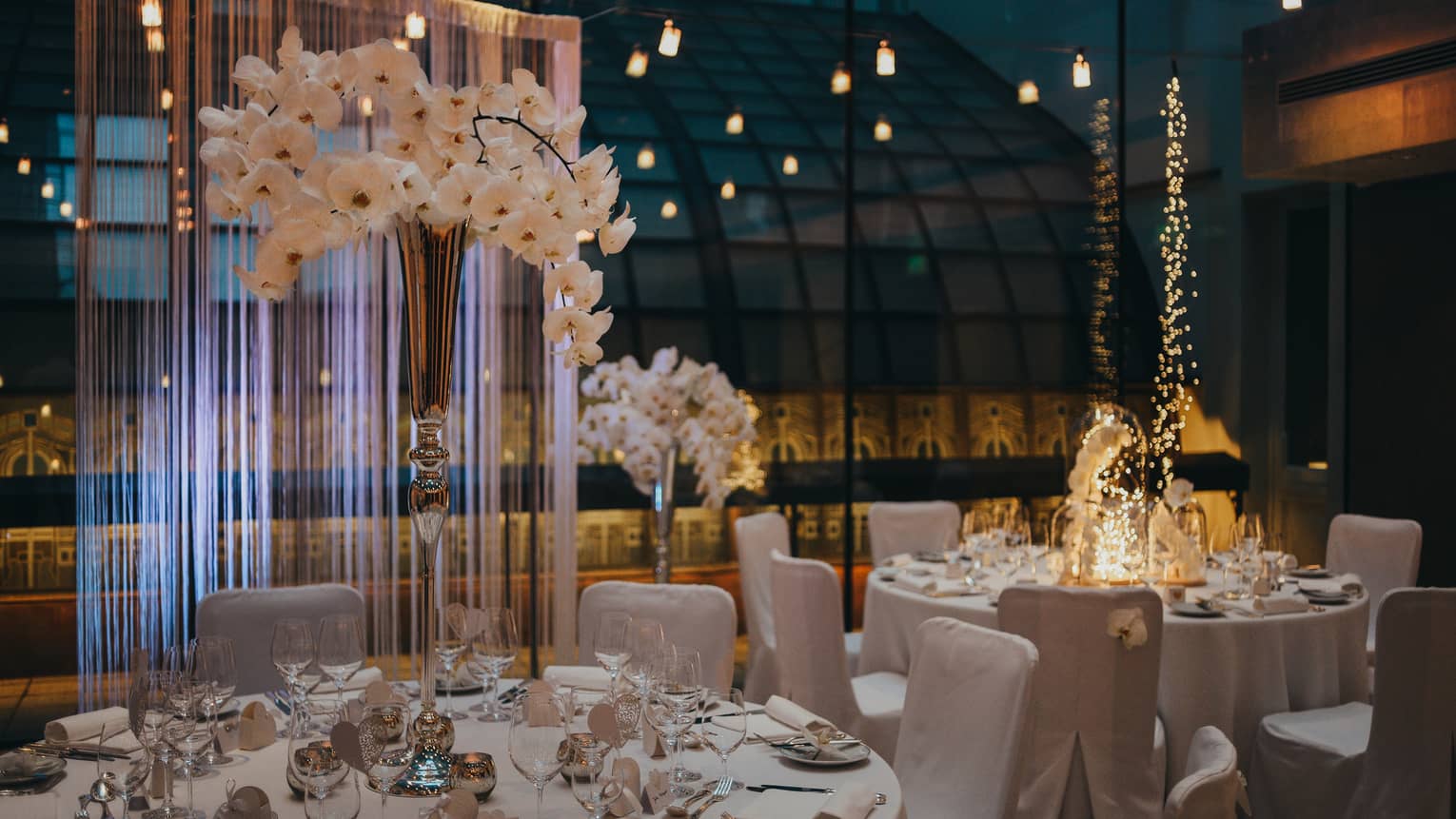 White dining tables and chairs with vase of orchids beside glass wall and reflecting lights