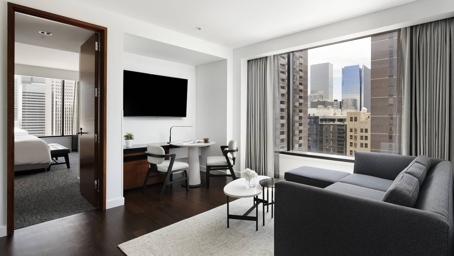 A hotel suite with a table and chair under a tv, a black sofa with a coffee table, and a bed and bench in a bedroom.