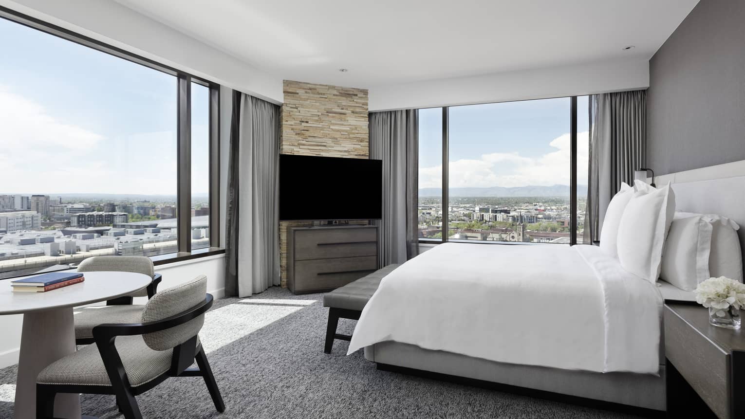 A hotel room with a white sheet covered bed, a TV, small white table with grey chair and large windows on two walls.