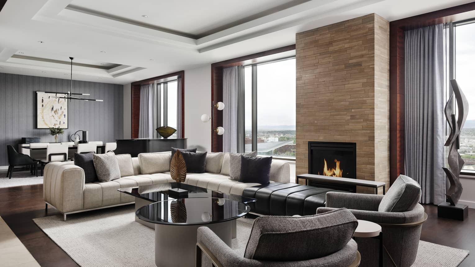A lounge seating area with a fire place, a white sofa, two grey chairs, and a black table between them; there are also large windows.