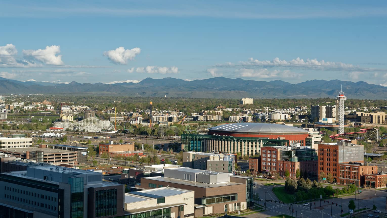 Aerial city view featuring an amusement park, a large round building, a TV tower, mountains in the distance under blue sky.