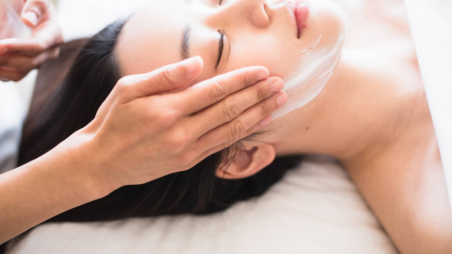 Close-up of spa attendant's hands rubbing white cream on woman's face as she lies on massage table