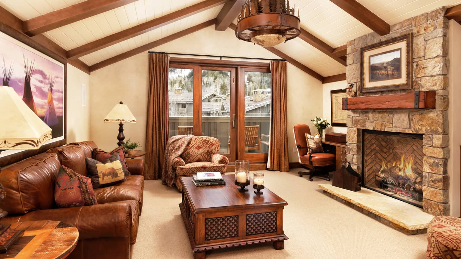 Rustic living room with leather sofa, armchair, tables, desk, stone fireplace, vaulted wood-beam ceiling and balcony doors with mountain view