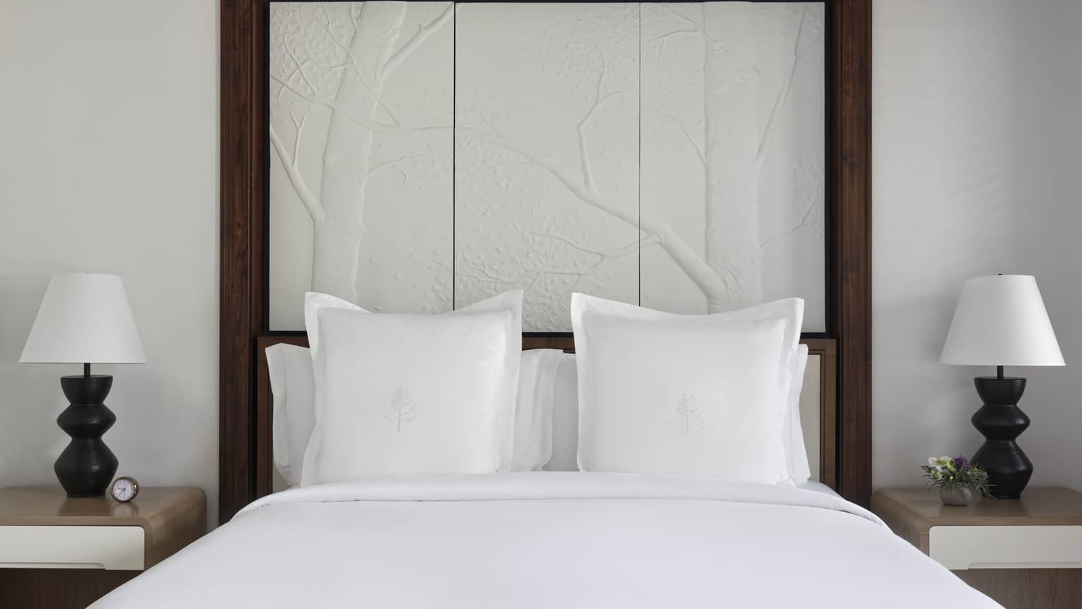 King bed with white linens and pillows with the Four Seasons logo, set against a tree branch headboard and flanked by nightstands with lamps