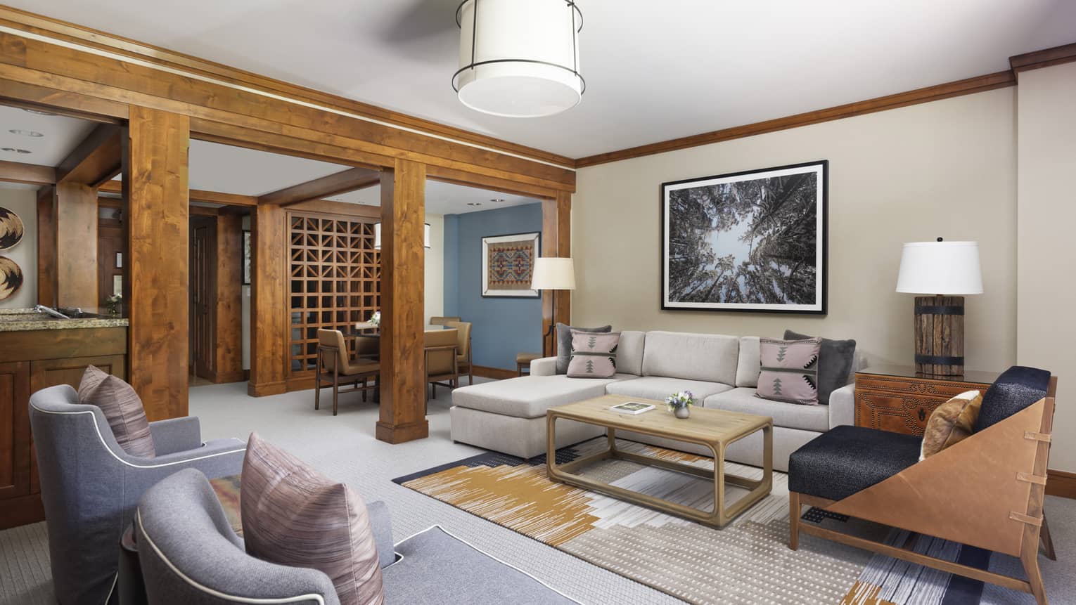 Luxury resort living room with a grey sectional, armchairs, coffee table, forest-themed wall art, and wooden beams separating the dining area