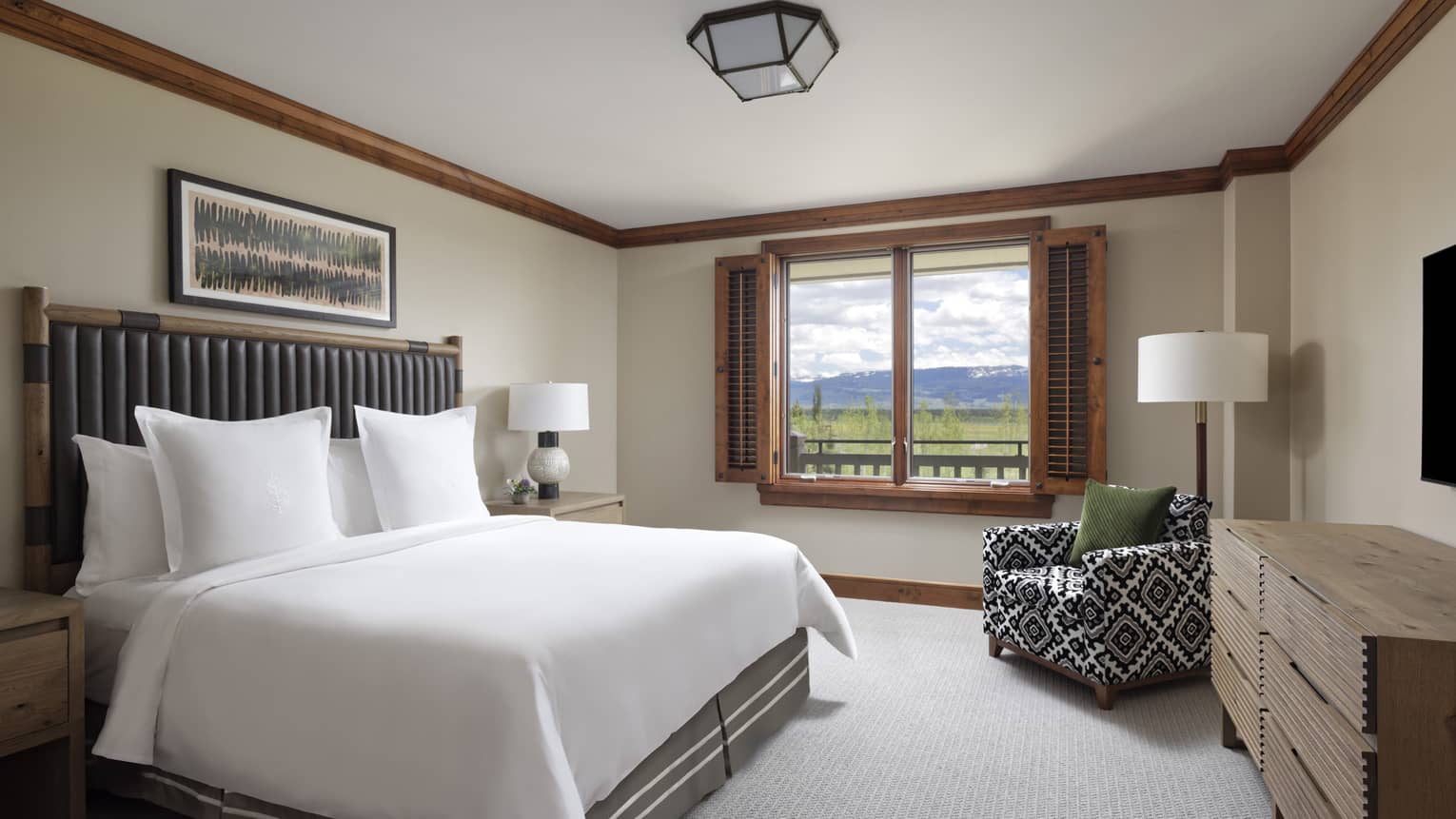 Bedroom with a white bed, dark headboard, patterned armchair, dresser, TV and a window with a mountain view