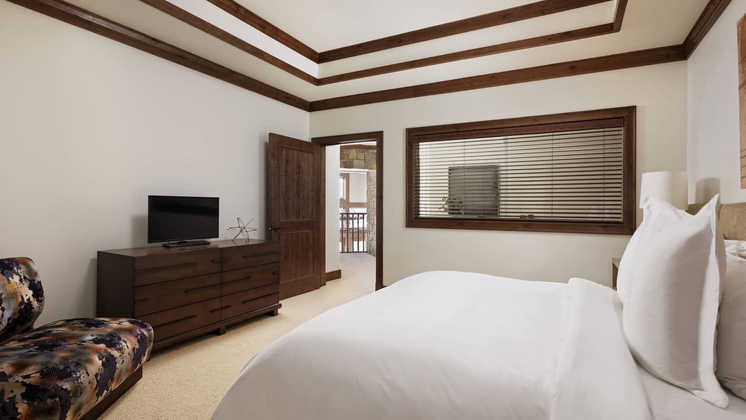 Bedroom with a white bed, dark wood dresser, small TV and patterned armchair, under a coffered ceiling with wood beams