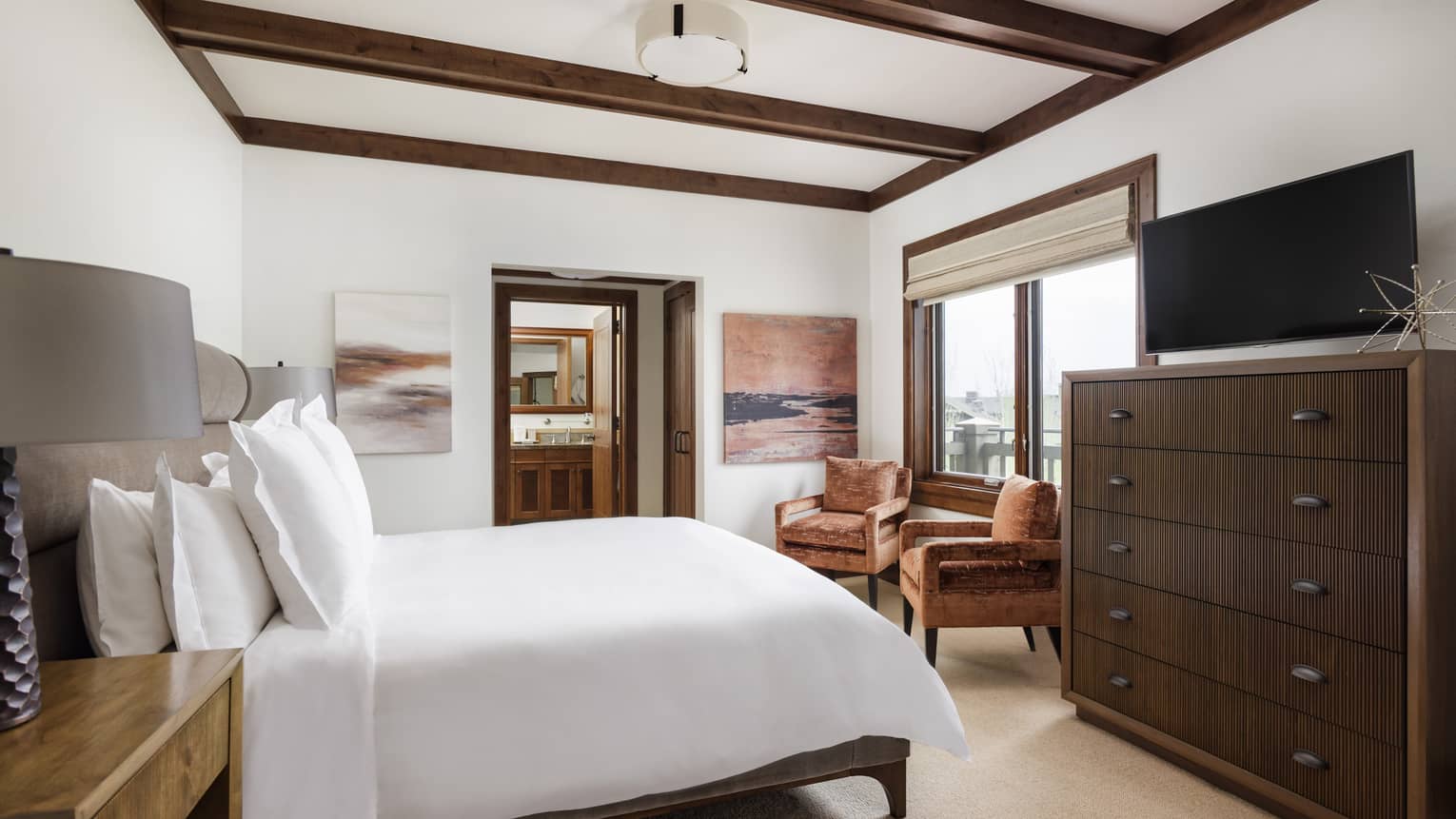 Bedroom with a king bed, dark wood dresser with TV, large windows, two armchairs and wall art, connected to a bathroom through an open doorway