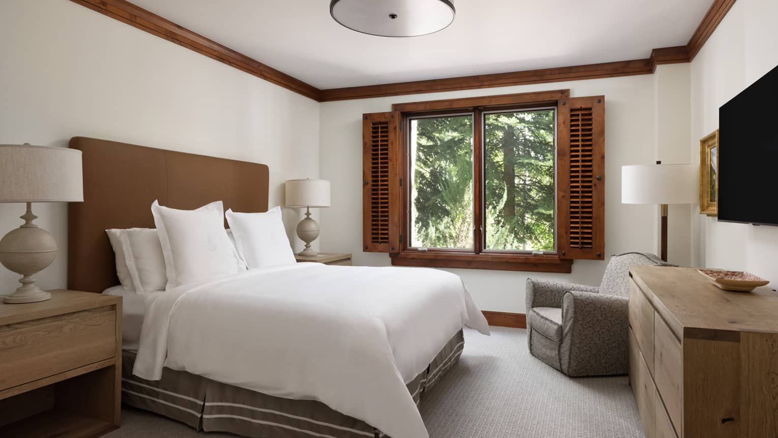 Bedroom with a king bed, brown headboard, side tables with lamps, dresser, armchair, TV, and window with wooden shutters overlooking trees