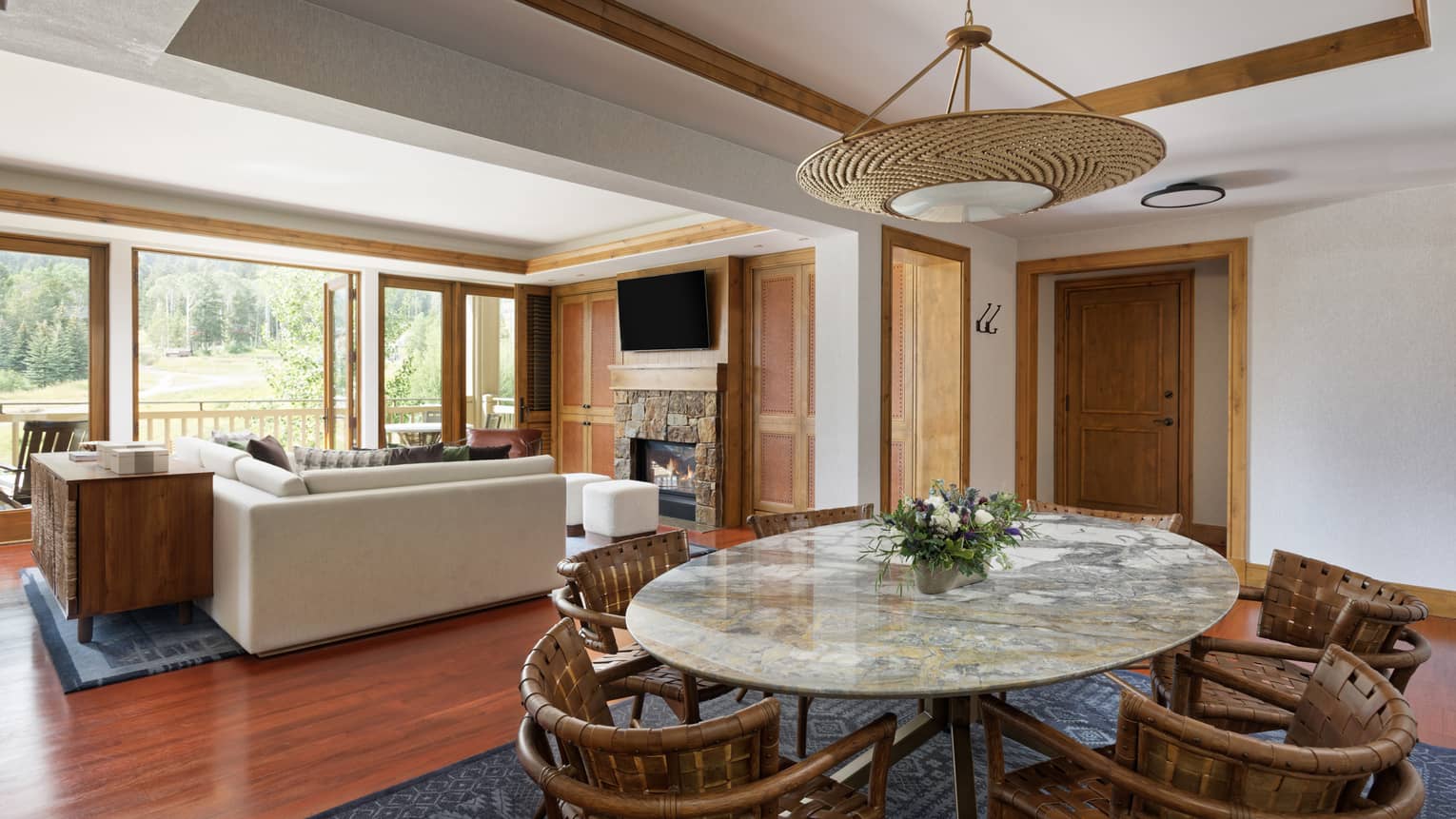 Luxury suite with a marble dining table, woven chairs, sectional sofa, stone fireplace, TV and large windows opening to a scenic balcony view