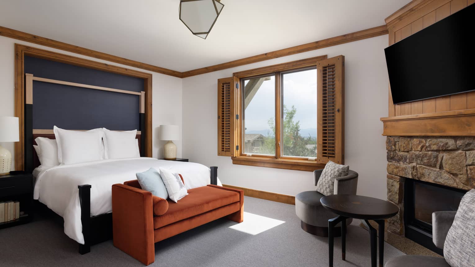 Luxury resort bedroom with a king bed, burnt orange chaise, grey armchairs, stone fireplace, TV and window with wooden shutters