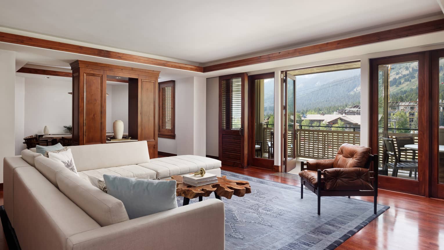 Suite living room with a cream sectional sofa, rustic wood coffee table, leather armchair and large doors opening to a balcony with a mountain view