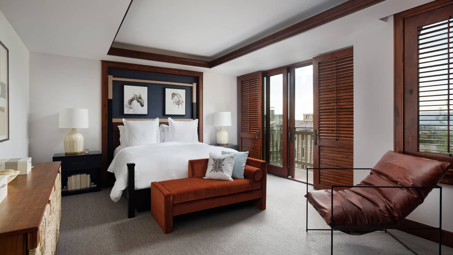 Luxury resort bedroom with a king bed, nightstands with lamps, burnt orange chaise, leather lounge chair and dark wooden doors opening to a balcony view