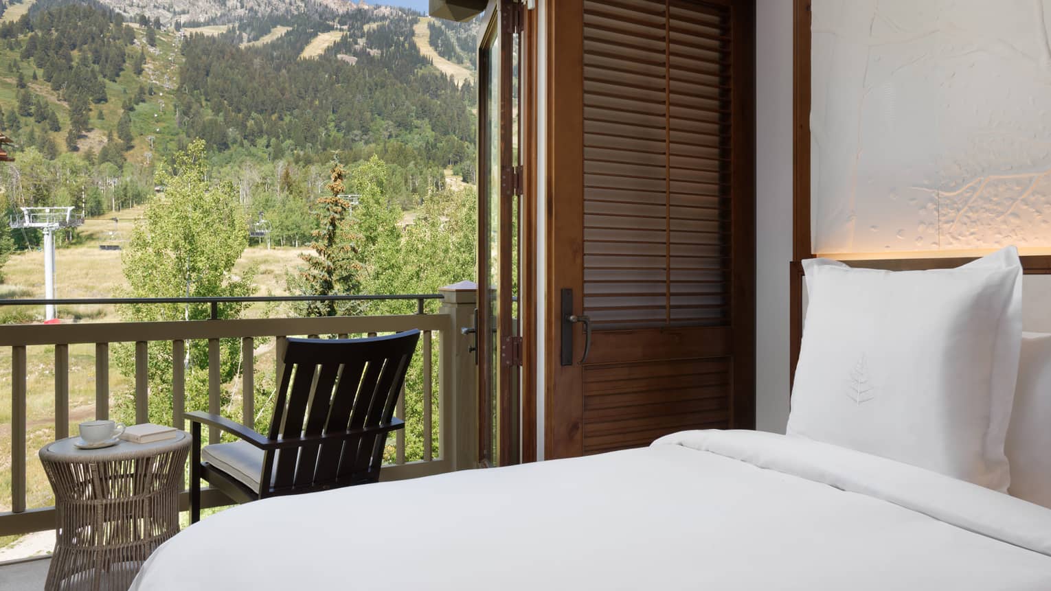 Bedroom with a white bed facing a balcony with mountain views, featuring wood accents, a lounge chair, and a small side table with a cup and book