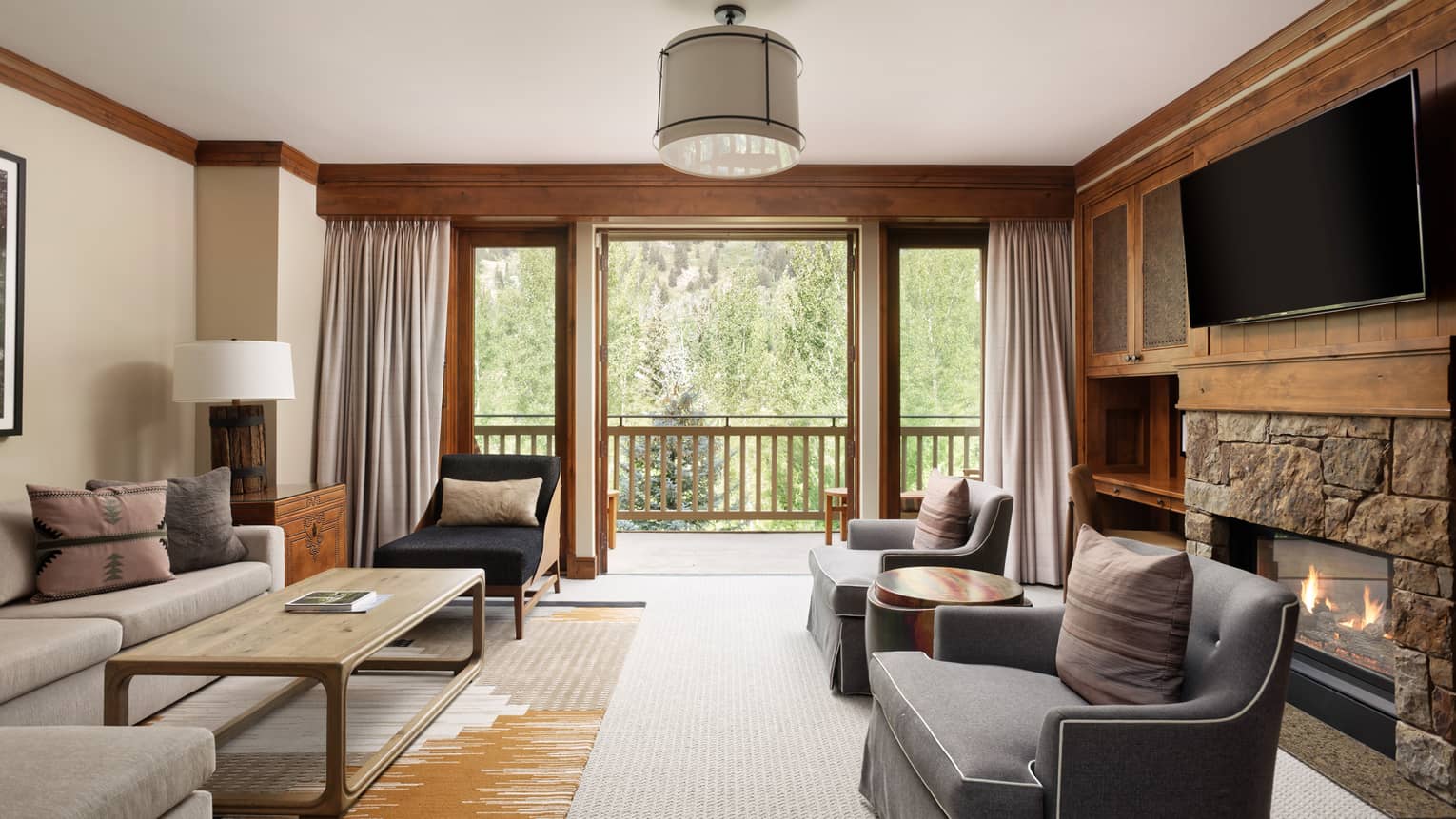 Luxury living room with stone fireplace, grey armchairs, large sofa, TV and walk-out balcony with forest view