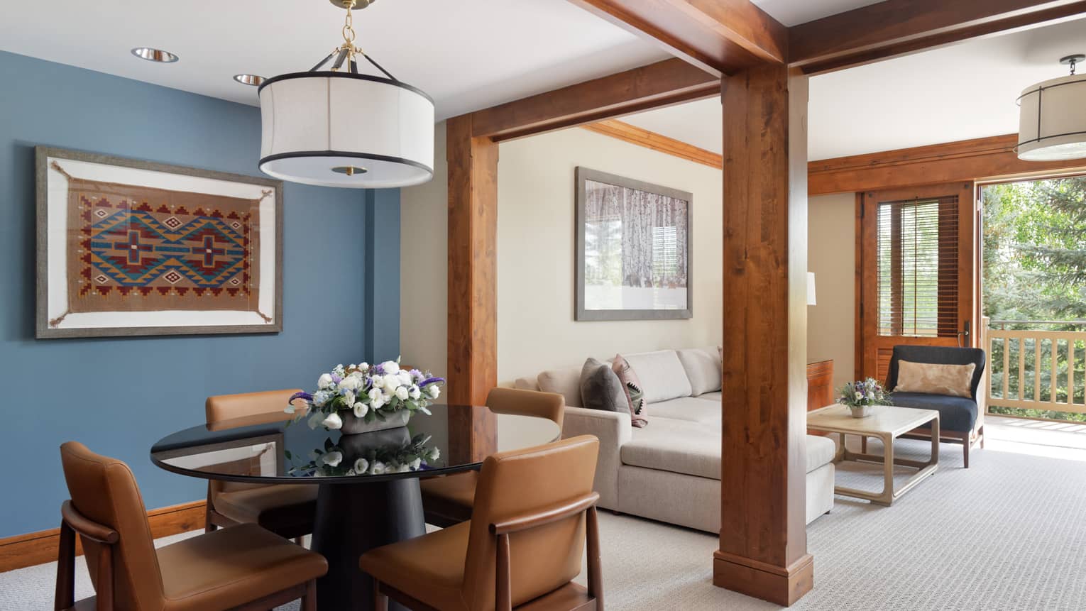 Dining area with a round glass table, leather chairs, and a blue accent wall displaying a framed textile art piece, opening to a living room with a sectional sofa