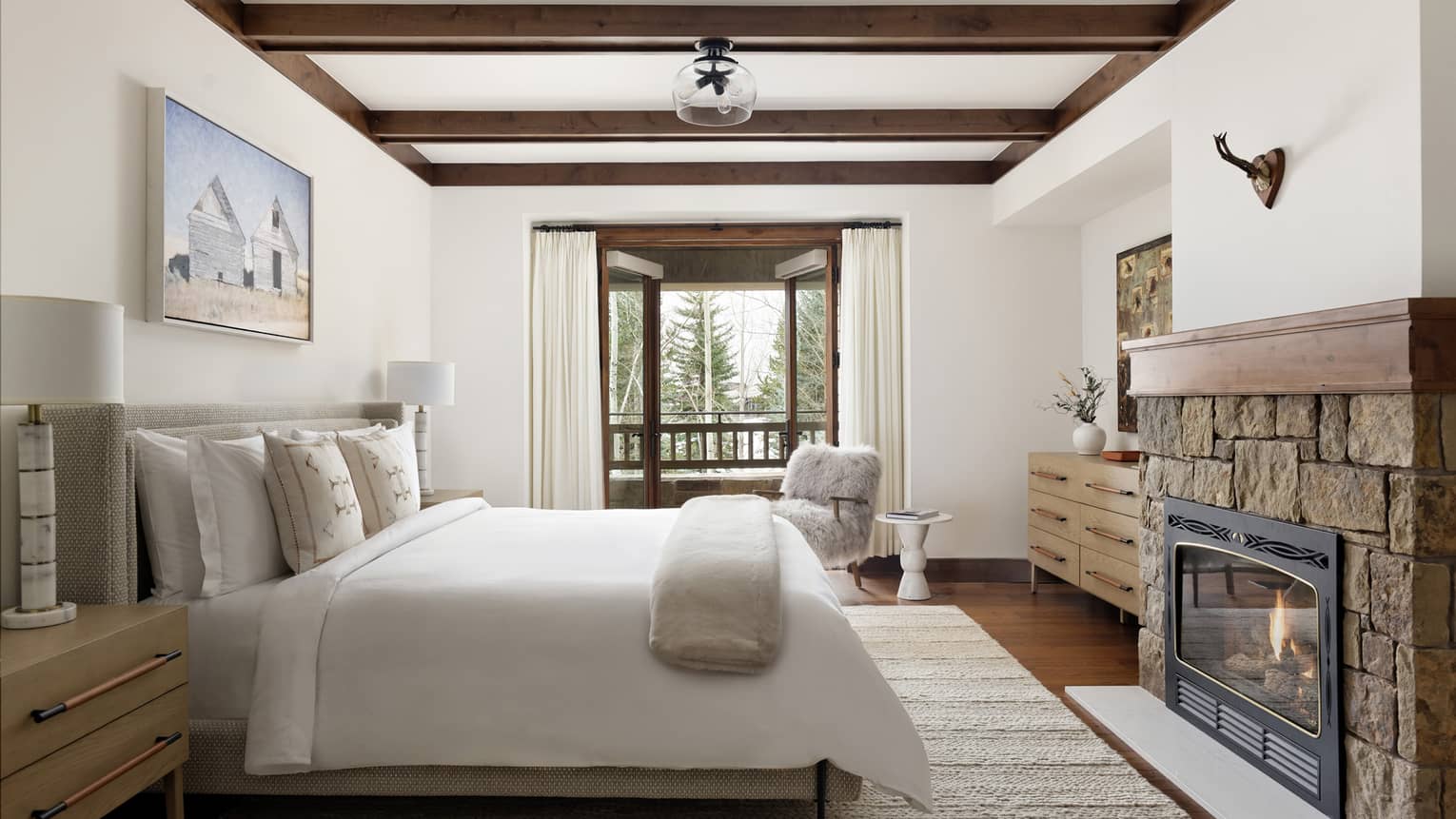 Bedroom with a king bed, nightstands with lamps, wall artwork, stone fireplace, dresser, fluffy armchair, and balcony 