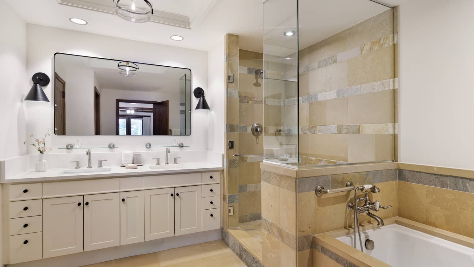 Luxury resort bathroom with double vanity, large mirror, black sconces, glass shower and stone-tiled bathtub