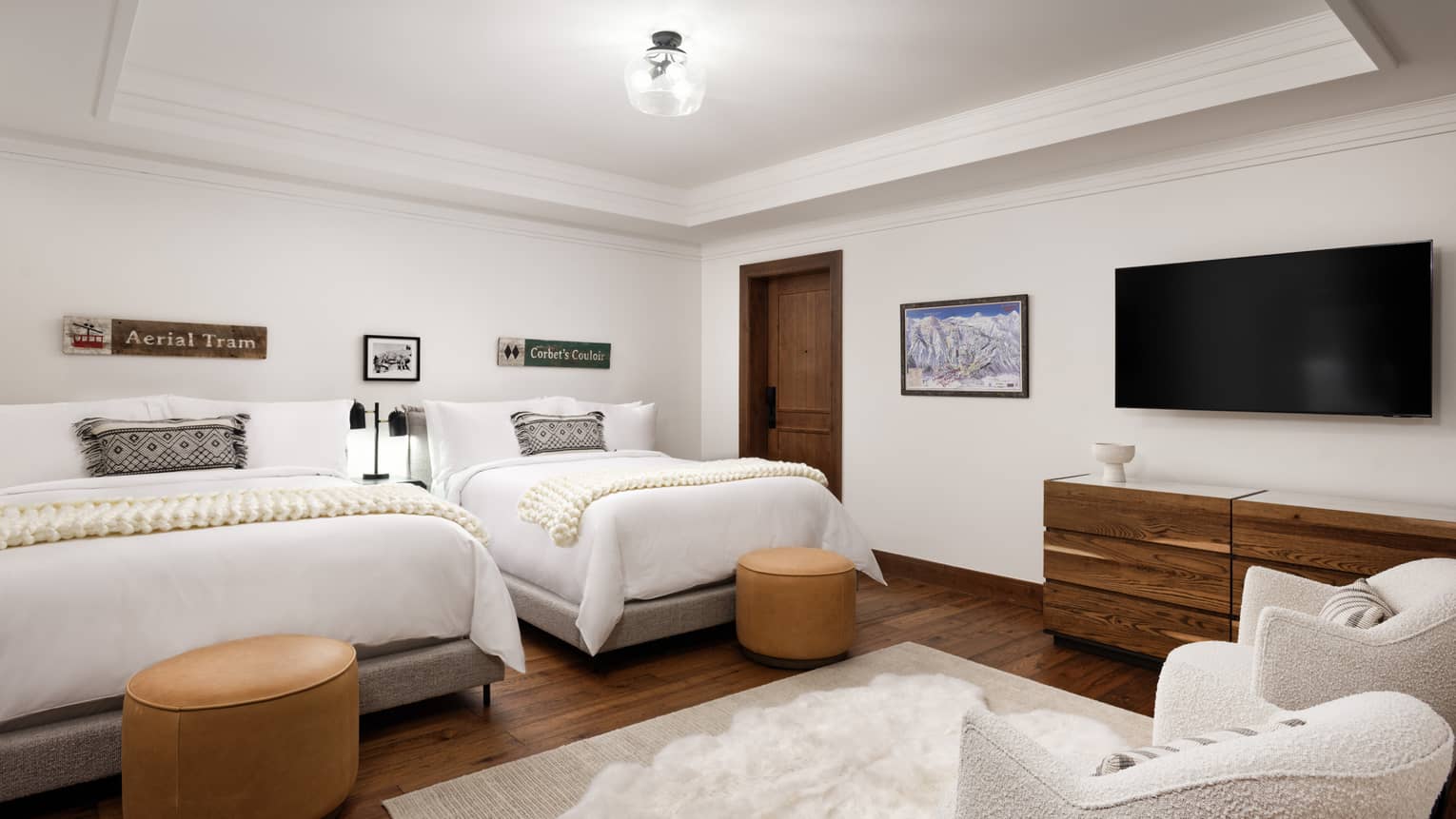 Bedroom with two beds, wooden dresser, wall art, TV, leather ottomans, and armchairs