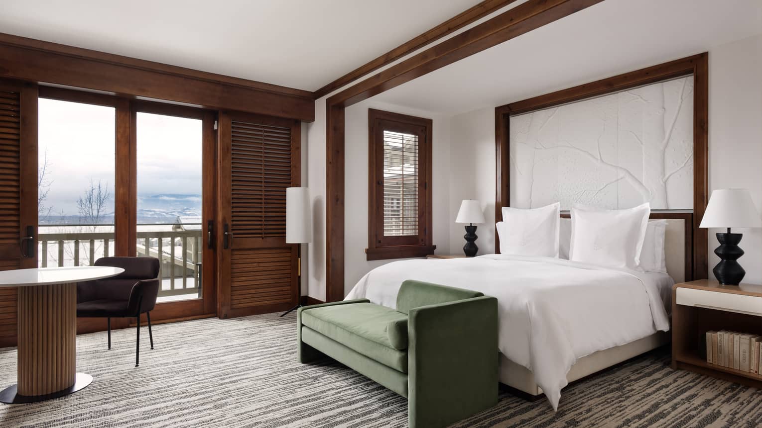 Stylish guest room with a white bed, dark green bench, table with chair, nightstands with lamps, wooden accents and large windows showing a snowy valley