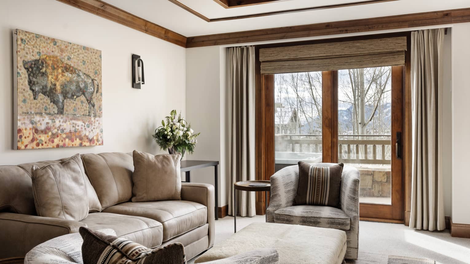 Luxury resort living area with a beige leather sofa, patterned armchairs, bison artwork on the wall, and mountain-view balcony