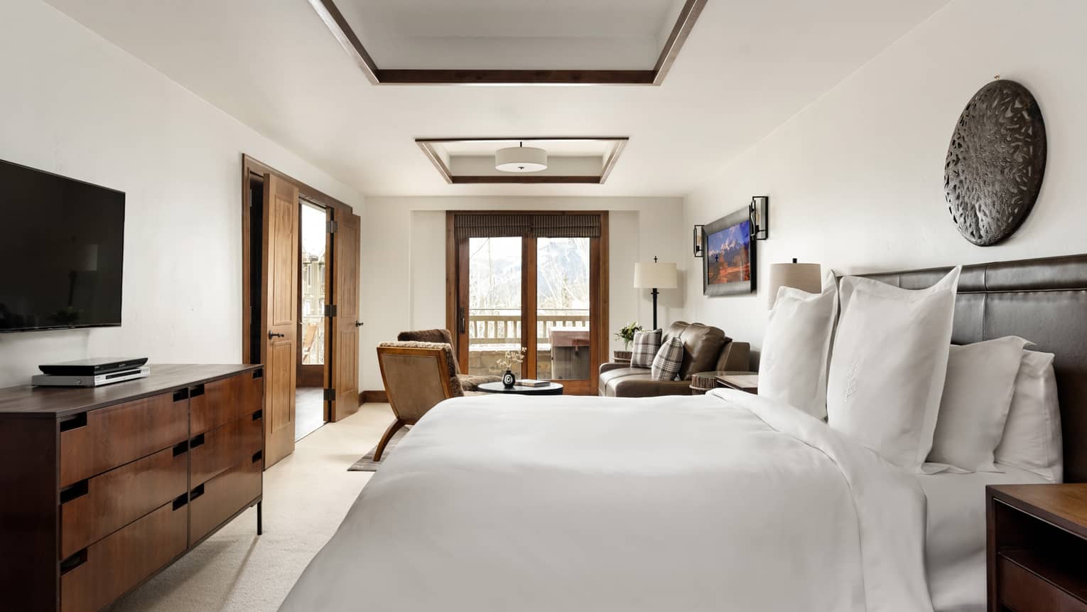 Bedroom with a king bed, wall-mounted TV, dresser and cozy seating area by wooden-framed doors leading to a balcony