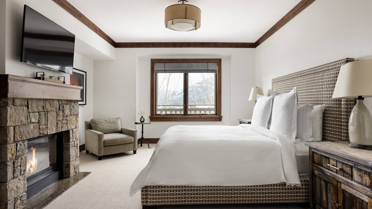 Bedroom with a patterned upholstered king bed, nightstands with lamps, stone fireplace, TV, and a cozy armchair near a window with mountain view