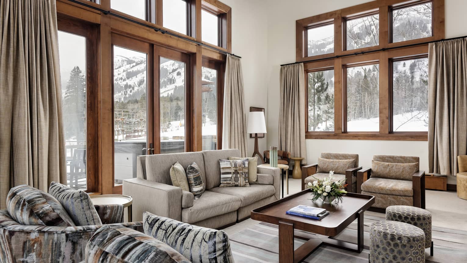 Luxury resort living room with tall windows, beige sofa, armchairs, ottomans, table and snowy forest view 