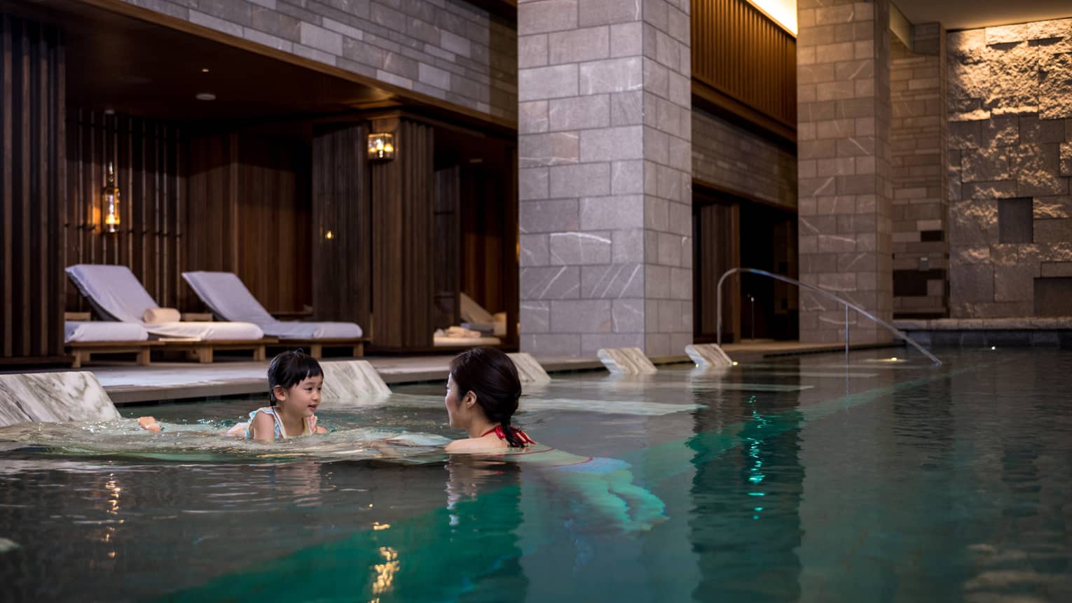 Woman leads young child into water in indoor swimming pool by marble in-water lounge chairs
