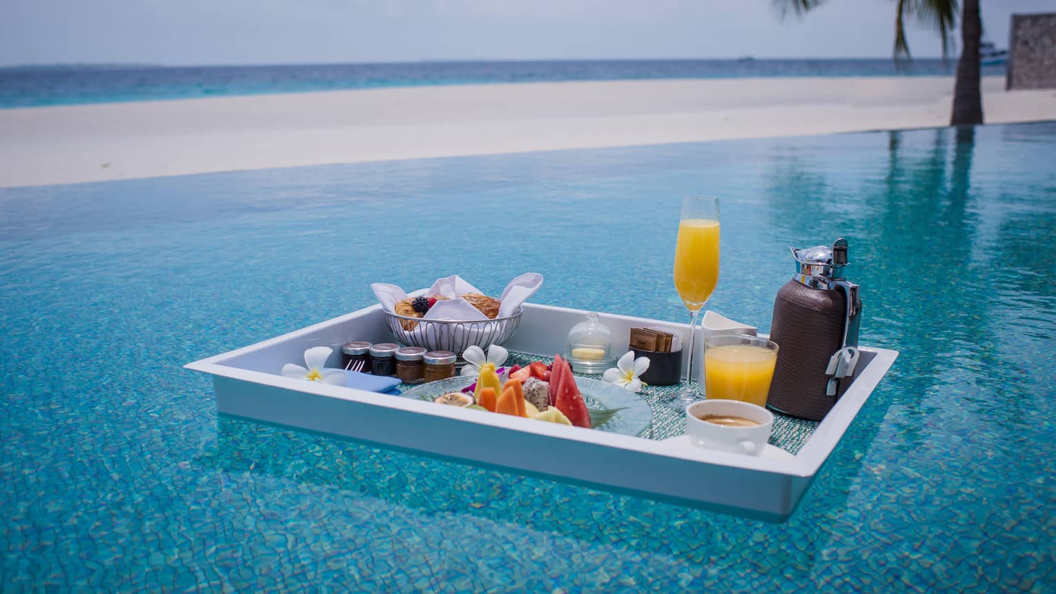 Floating in a shimmering turquoise pool, is a white tray with a selection of fruit, pastries, jams, beverages and flowers.