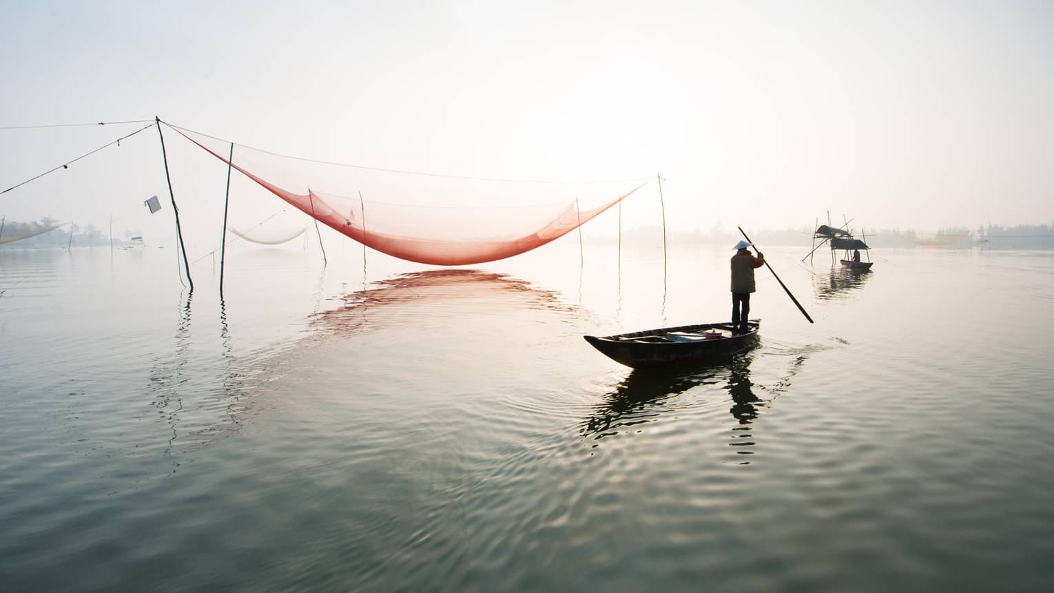 A man fishes standing up in a canoe as the sun shines brightly behind him. 