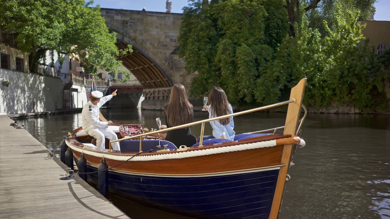 Small boat parked at dock on river, captain in white uniform points to bridge as two women drink Champagne