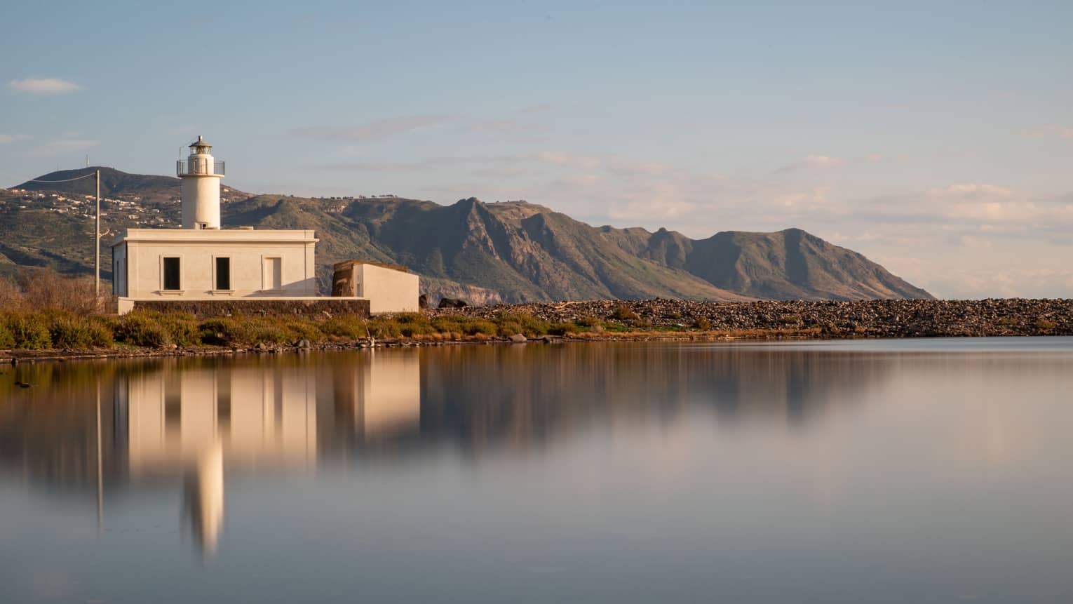 Short all-white lighthouse, reflecting on smooth water, mountains and blue sky in backdrop