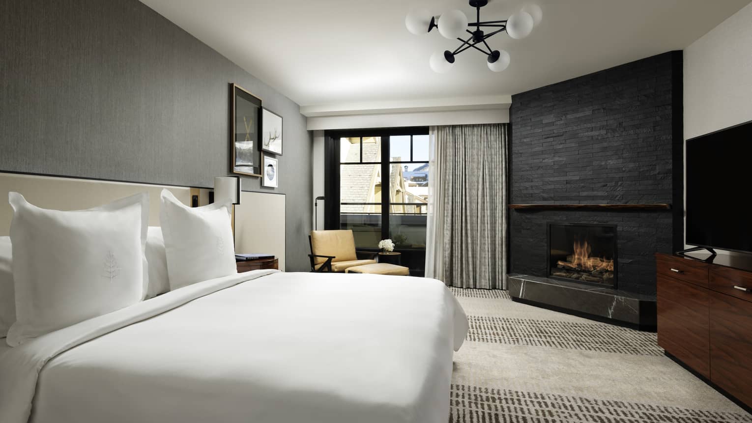 One-bedroom suite with King bed, corner fireplace, floor-to-ceiling window with grey curtains, yellow chaise