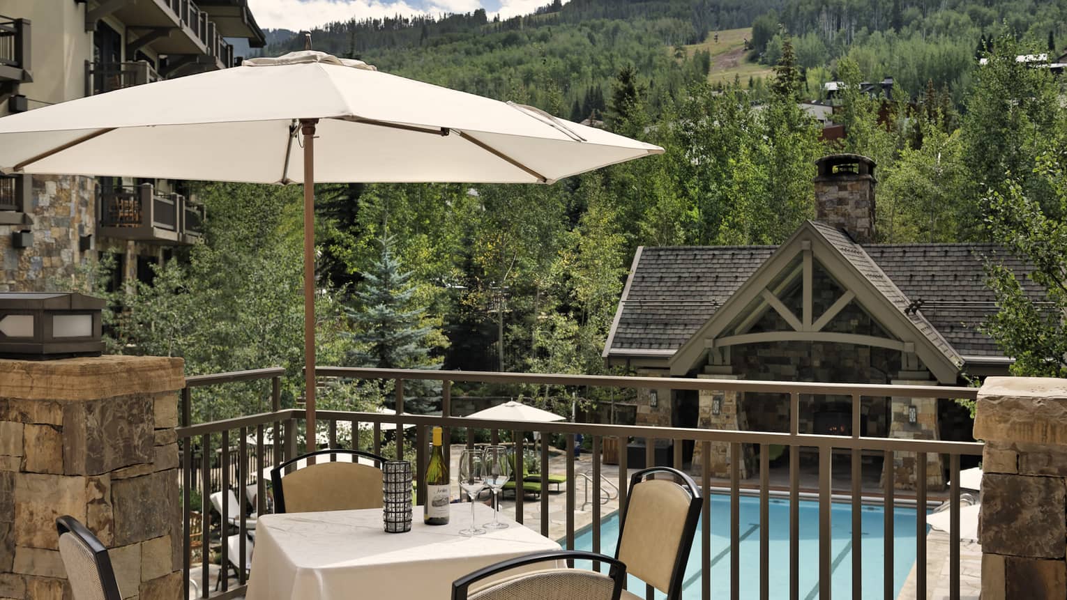 A balcony with a table and umbrella overlooking a pool and a cabin-like building, a forest of trees can be seen in the distance.