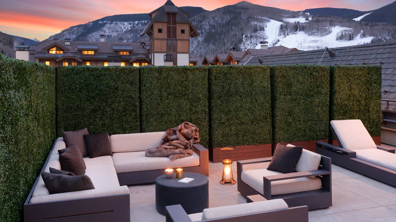 An outdoor seating area with. candles and white sofas near a cabin like village and mountains.