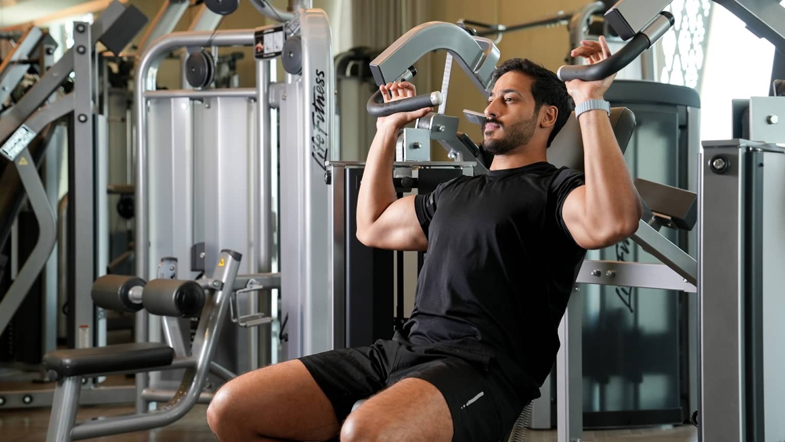 Man wearing black t-shirt and shorts works out on an arm weight machine at the gym