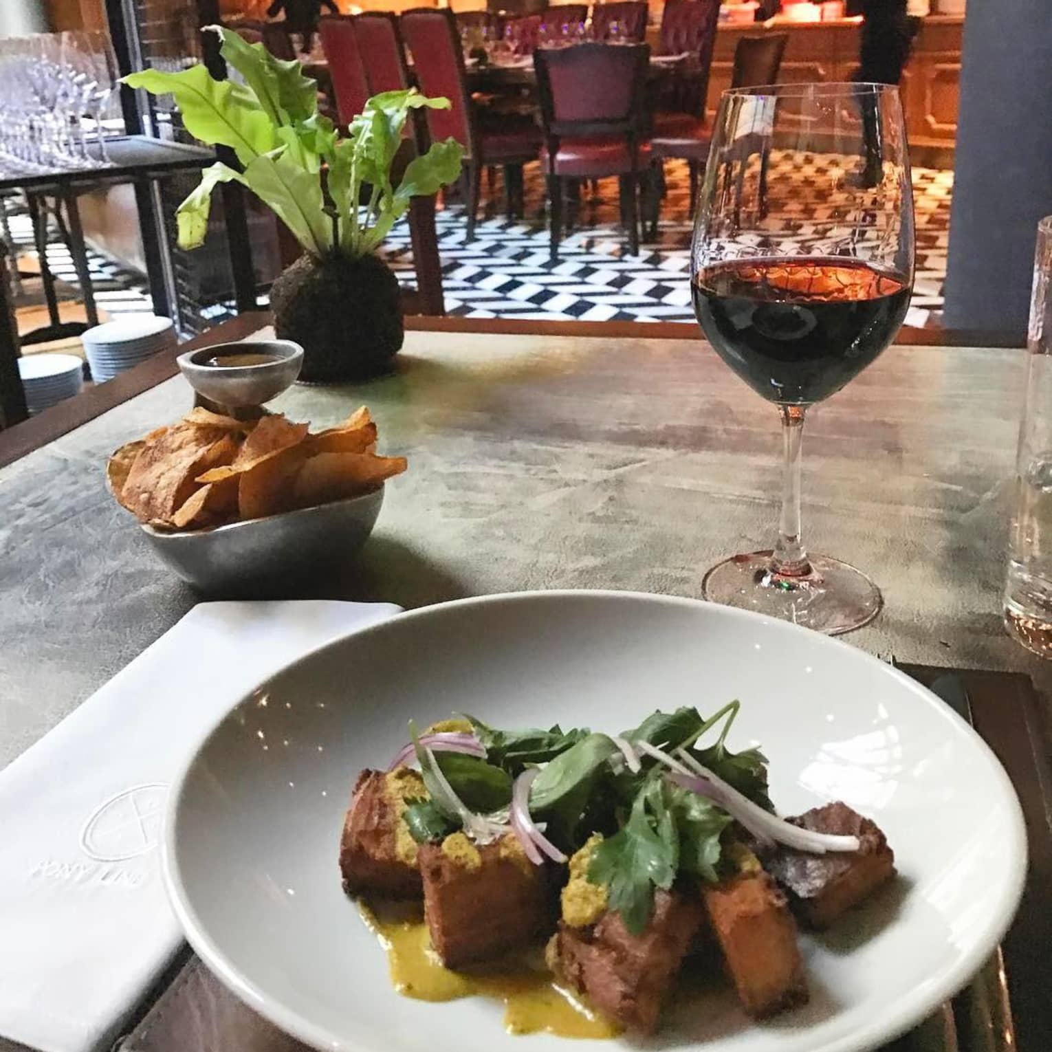 Meat dish with lettuce, sliced red onion, glass of red wine and bowl of crisps on restaurant table
