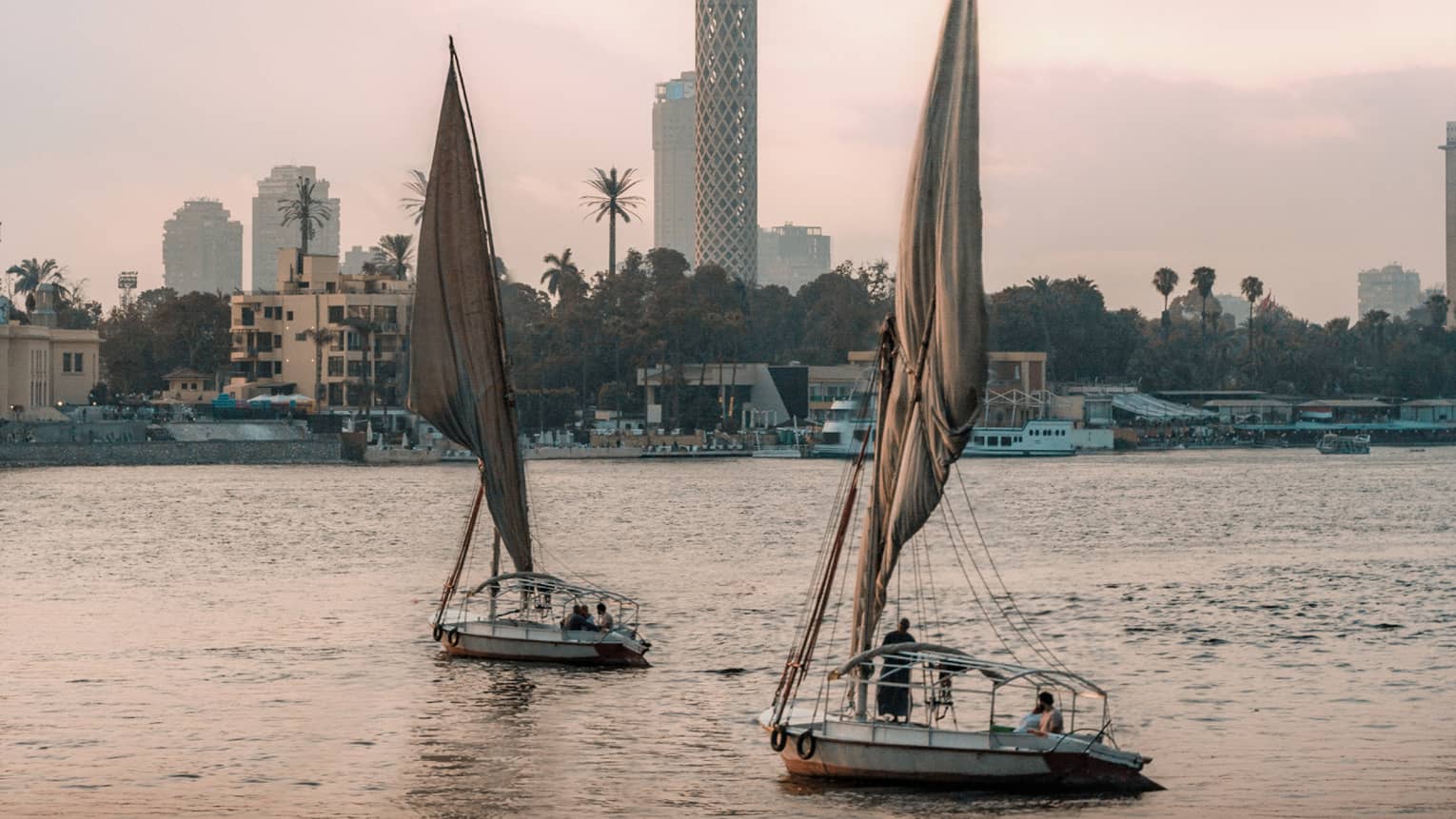 Two felucca boats drift on the water in the evening light, a busy shoreline and Cairo Tower in the distance. 
