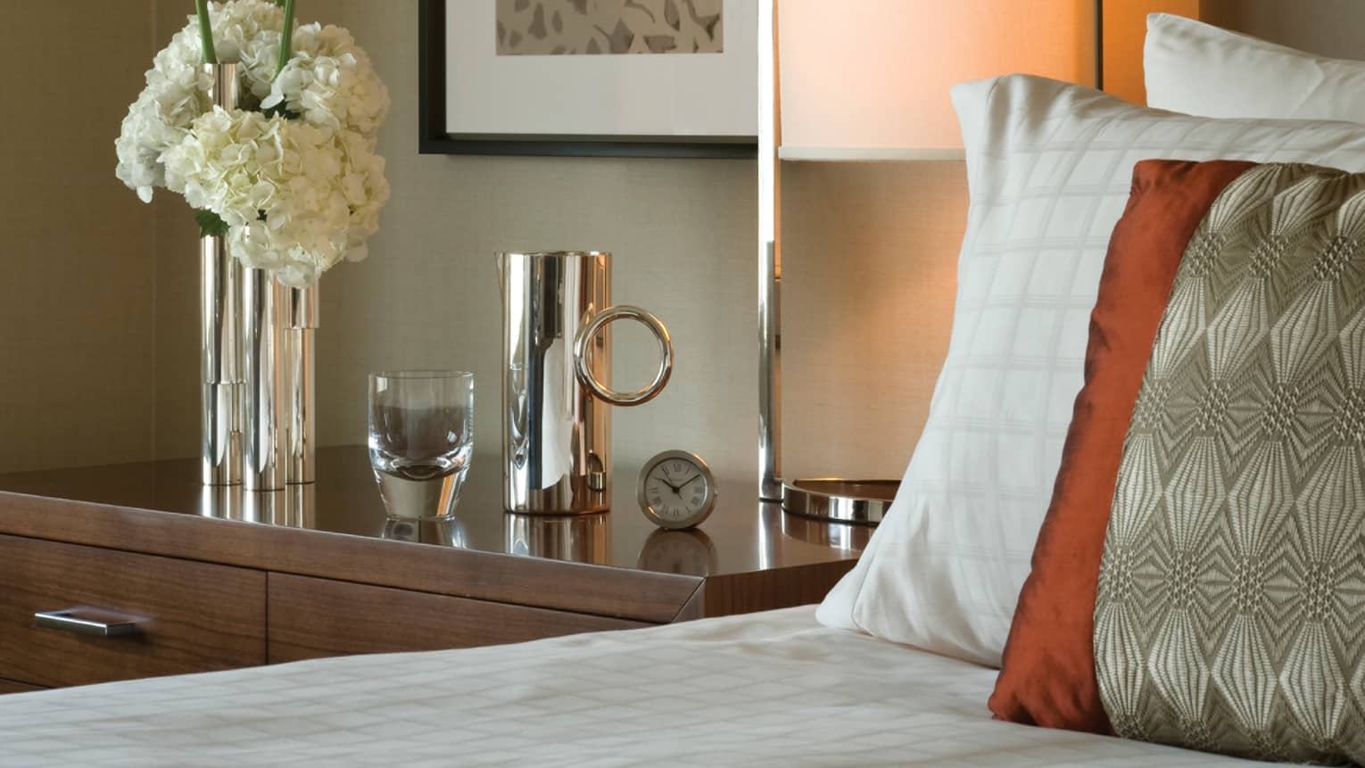 Premium Room close-up of bedside table with clock, silver vase with fresh white flowers, glass, canister, silk pillow on bed