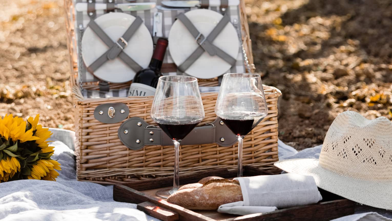 An open wicker picnic basket, two glasses of red wine and a baguette sit on a white blanket among rows of glowing vines.