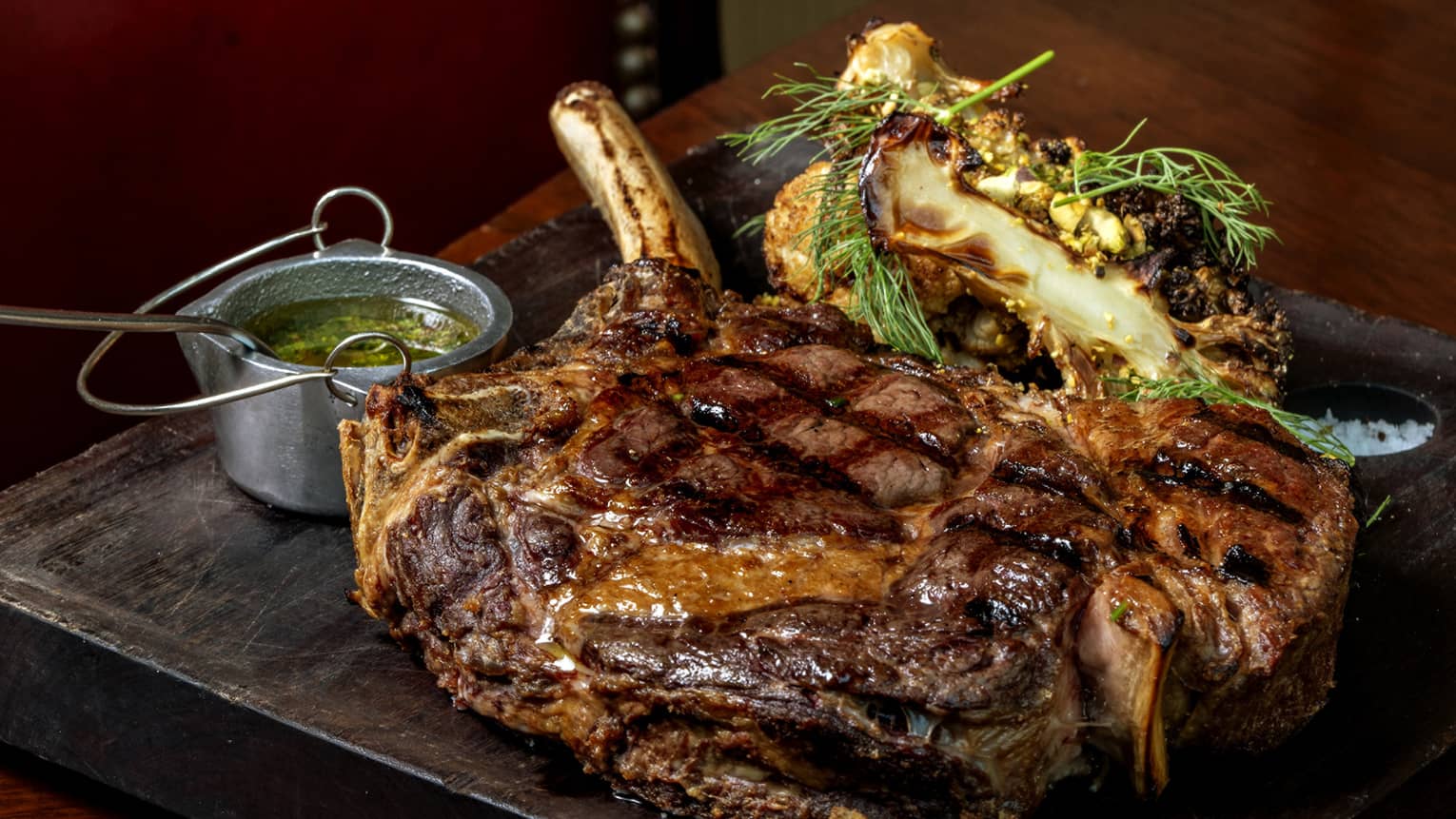 A thick, succulent cowboy ribeye steak rests on a wooden serving board, flanked by grilled vegetables and chimichurri sauce.