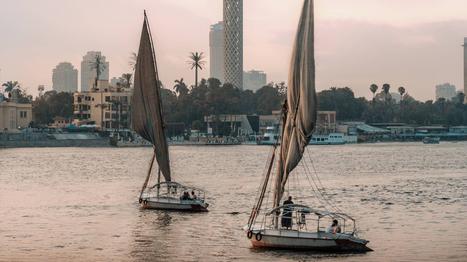 Two felucca boats drift on the water in the evening light, a busy shoreline and Cairo Tower in the distance. 