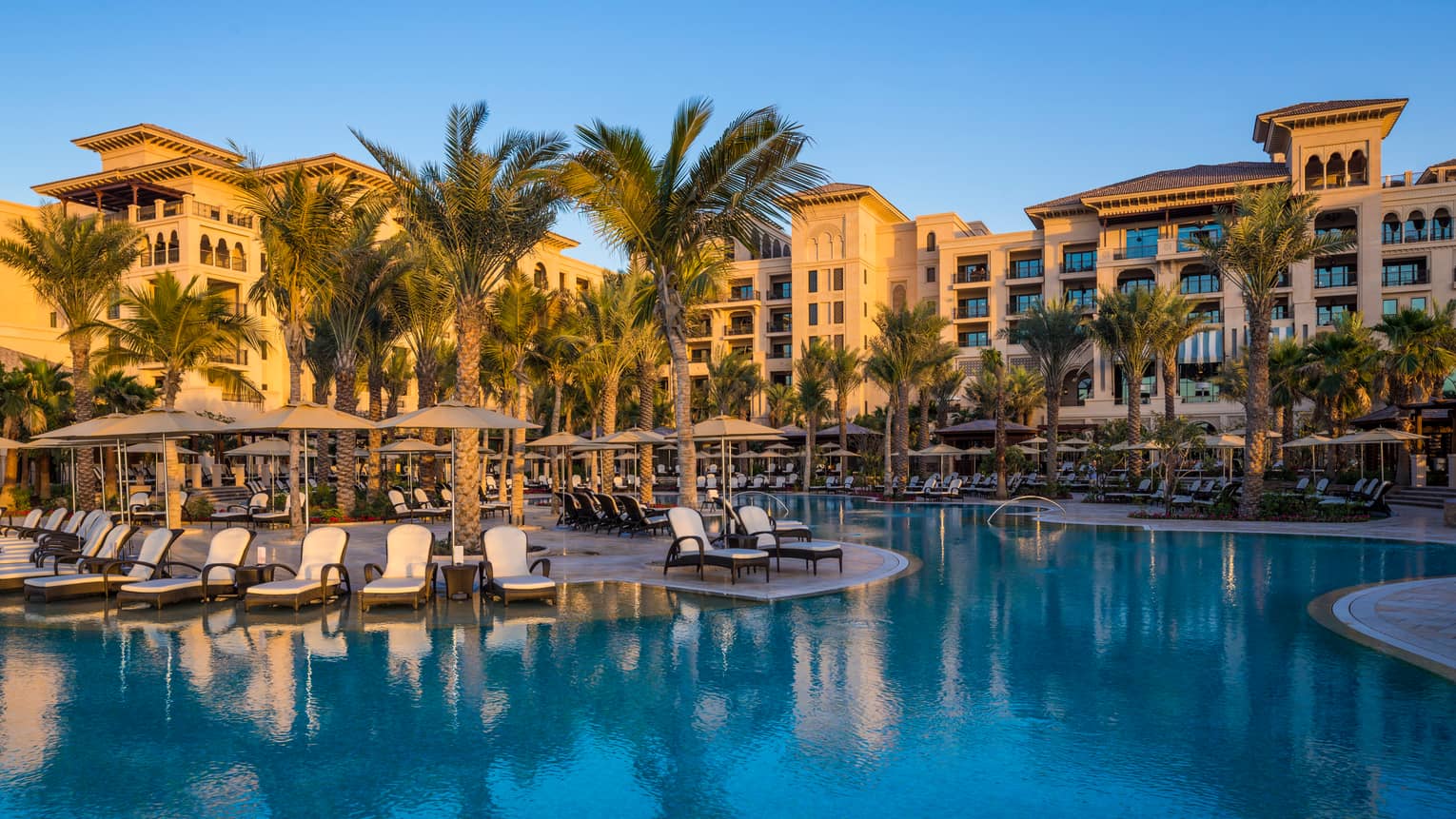Winding blue outdoor swimming pool lined with white chaise lounge patio chairs, palm trees