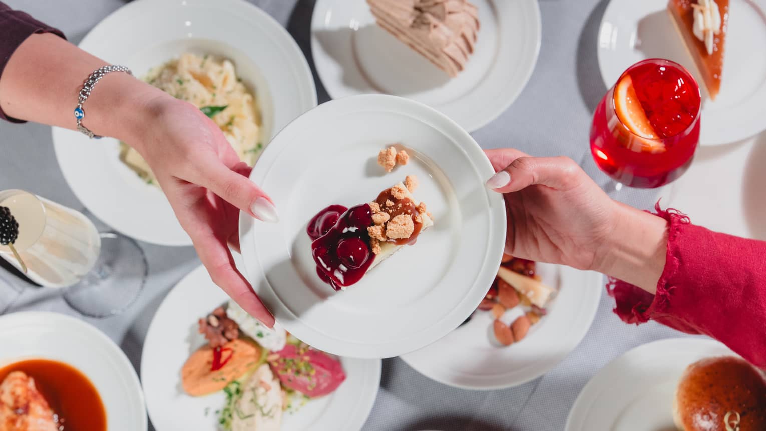 One hand passes along a white plate to another hand with a variety of dishes of food seen below on a table