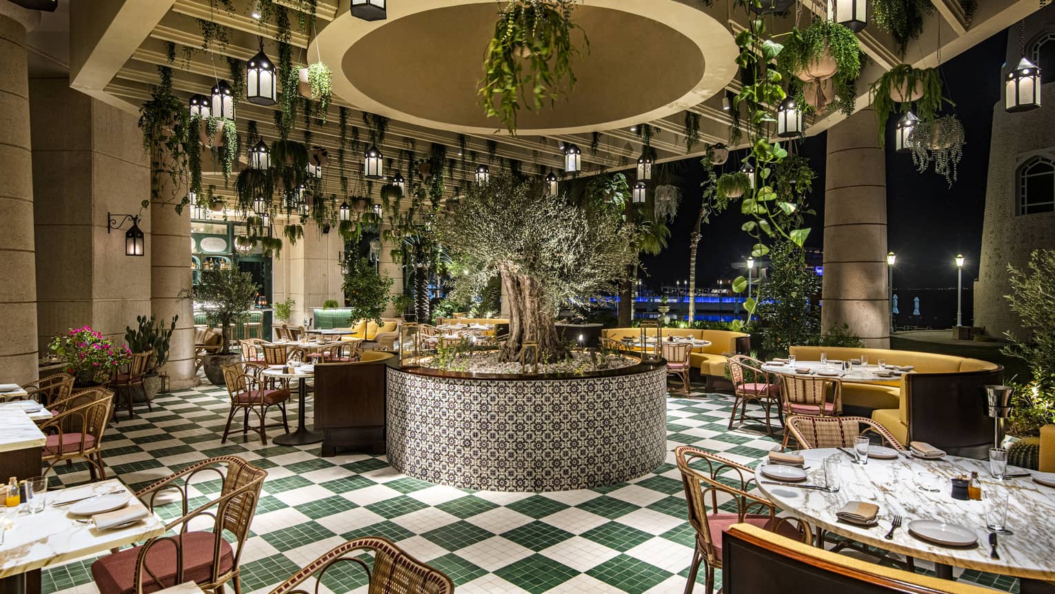 Curiosa restaurant interior featuring green-and-white checkerboard tile floors, rattan chairs, yellow banquets and plants and lanterns hanging from the ceiling