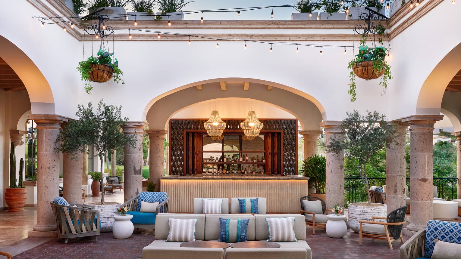 A spacious courtyard surrounded by arched openings. The space contains upholstered couches and chairs, along with potted and hanging plants, and string and pendant lighting.